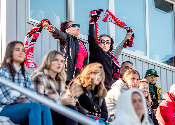 UHart alumni waving scarves and cheering on the Hawks at a soccer game during Hawktober weekend.