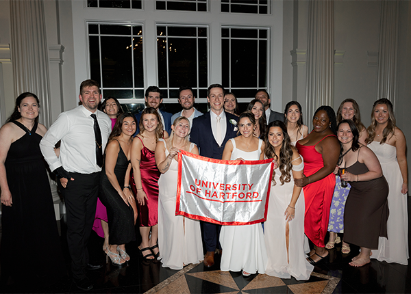 Danielle and her husband posing with a group of people and a University of Hartford banner.