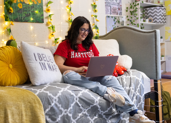 dorm room image with girl on her bed typing on laptop