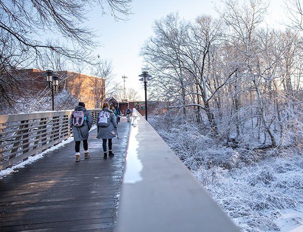 Students crossing the UHart bridge surrounded by trees covered with snow.